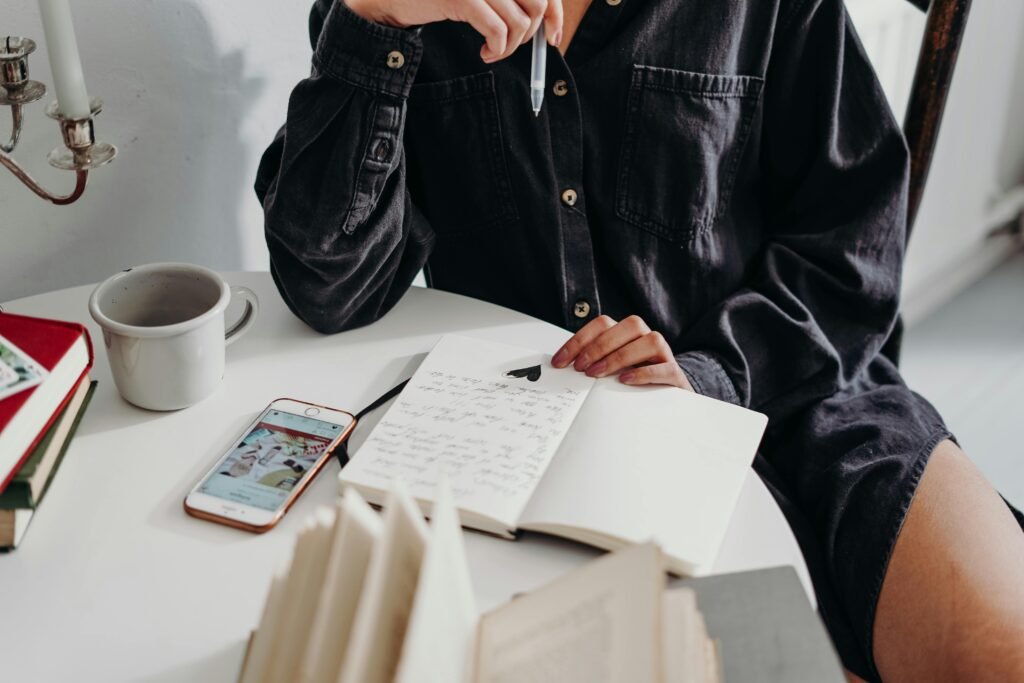 Woman seated indoors writing in a journal with coffee and a smartphone nearby.